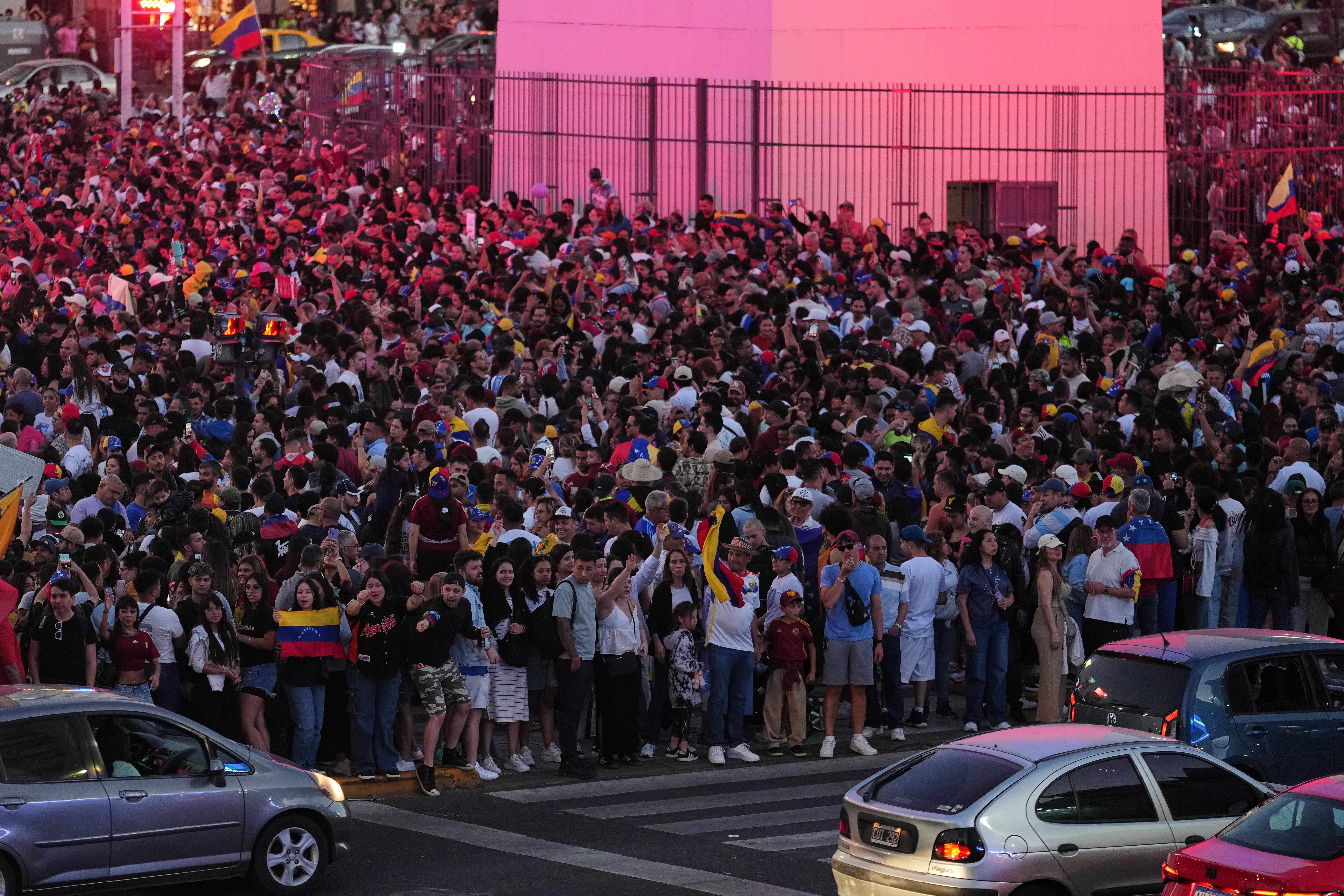 A large crowd gathers in a public square, mini holding Venezuelan flags.