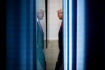 A color photograph of Donald Trump in profile, wearing a suit and red tie and looking at his reflection