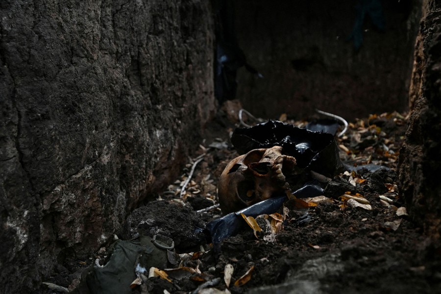 Part of a human skull rests at the bottom of a trench, surrounded by debris.