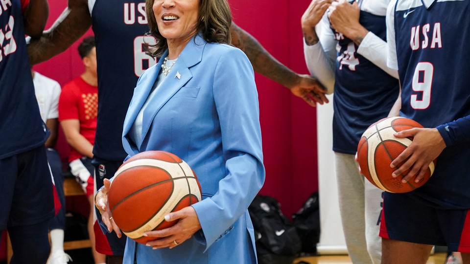 A photograph of Kamala Harris holding a basketball, with members of the American Olympic men's basketball team seen in the background