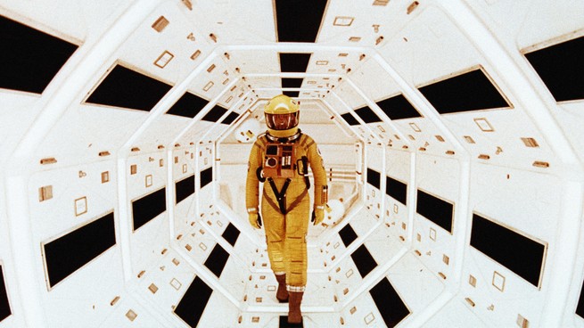 A man in a yellow spacesuit stands in a futuristic white tunnel in a scene from "2001: A Space Odyssey"