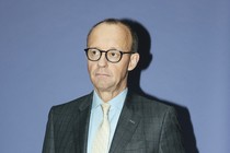 An unsmiling, vertical photo of Merz in a gray suit.