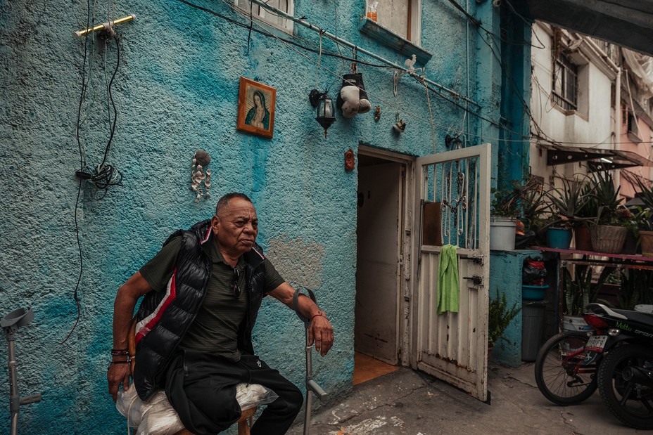 a man sits on a stool in an alleyway in front of a light blue concrete wall.