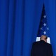 Color photograph of Donald Trump facing away from the camera, in front of the American flag and surrounded by a blue curtain.