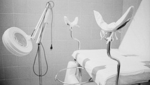 A black and white photograph of a patient examination room with stirrups and magnifying equipment