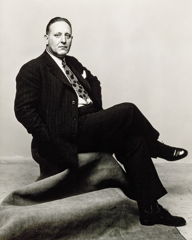 black and white photo of John Gunther wearing suit and tie, sitting in a chair, with hands in coat pockets and legs crossed
