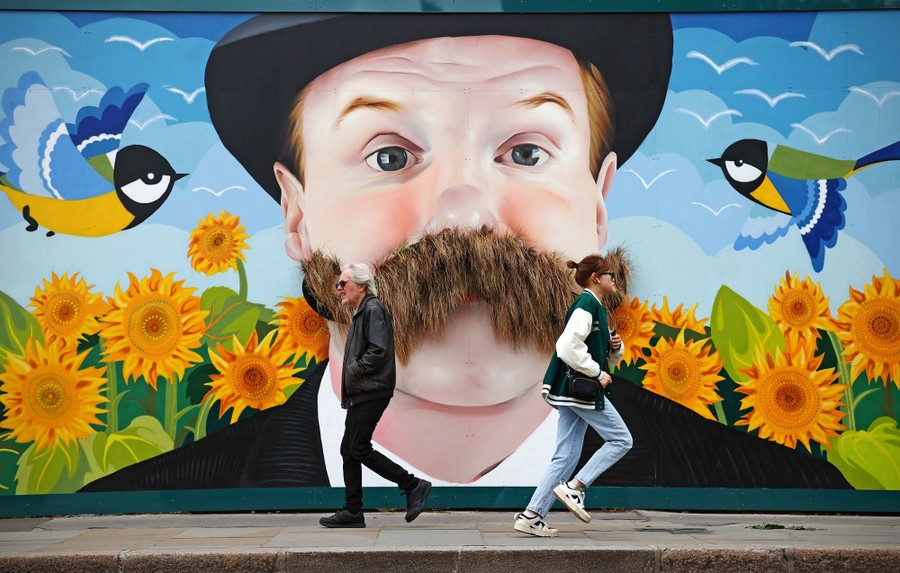 Two people walk past a large mural on a wall, depicting the face of a man with a bushy mustache.