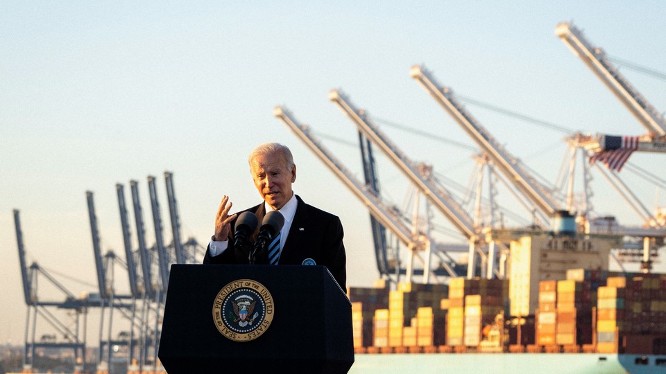 Joe Biden speaking from a lectern in a port, stacks of shipping containers visible on the background.