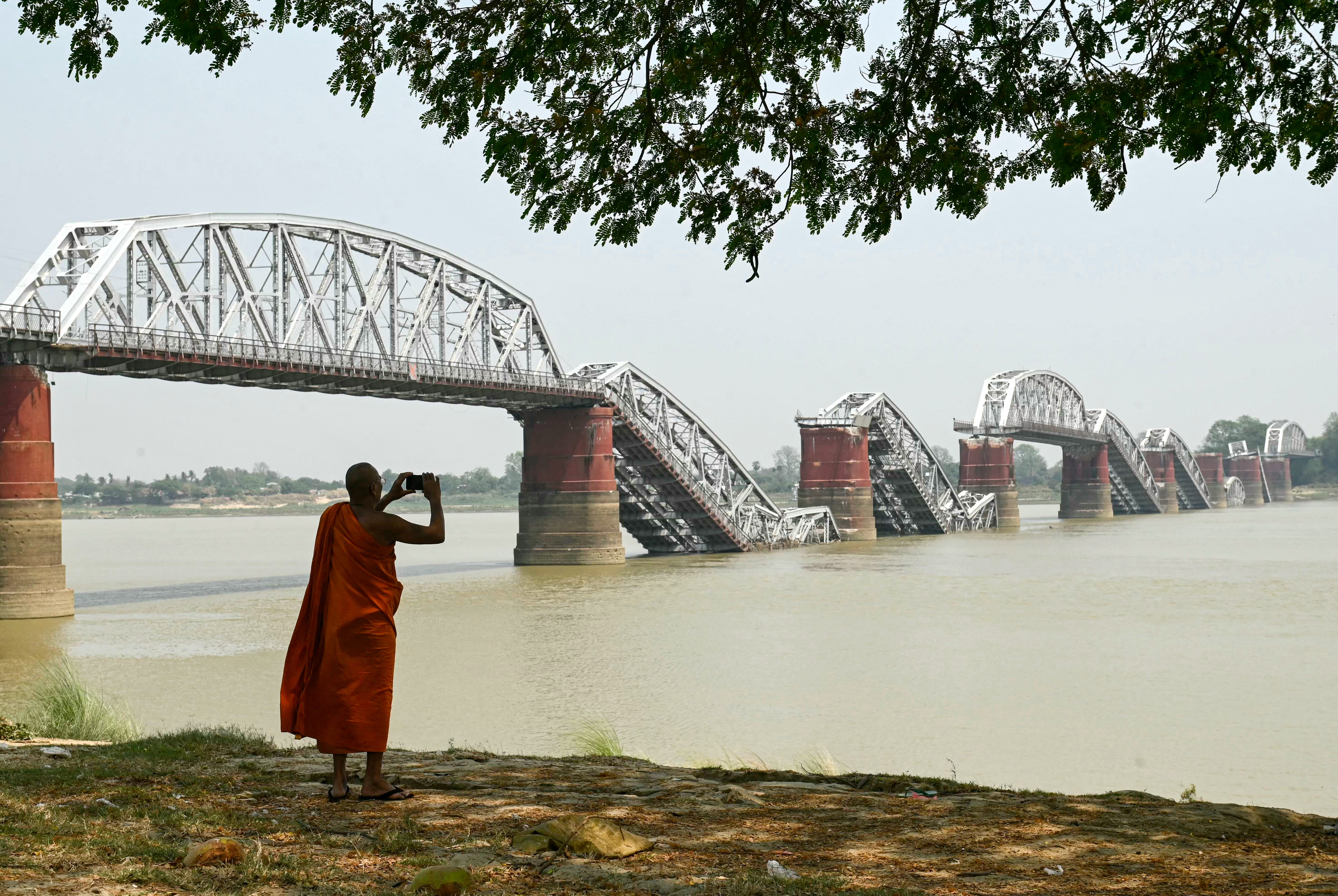 A monk takes photo on the banks of a river, looking toward a long bridge that has collapsed in several sections.
