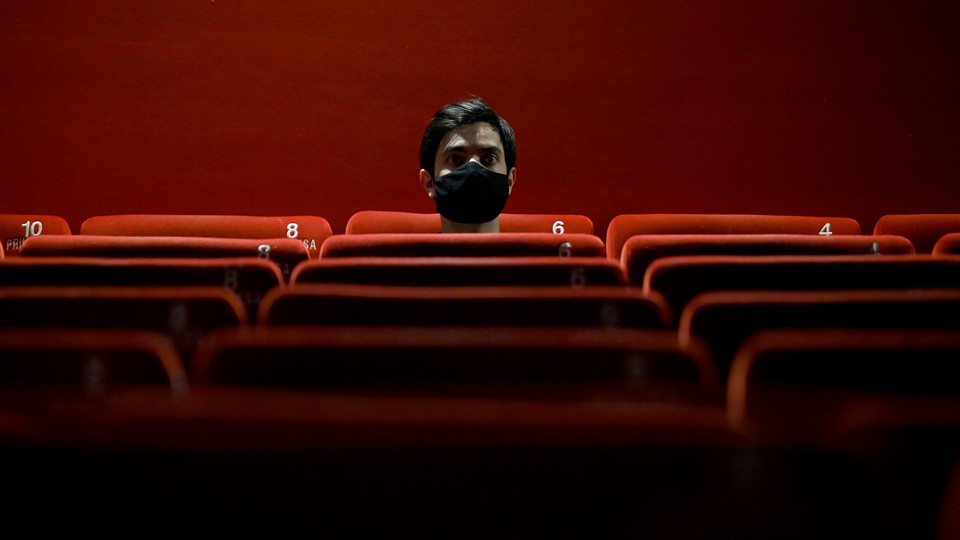 Person sitting alone in a red theater wearing a black mask