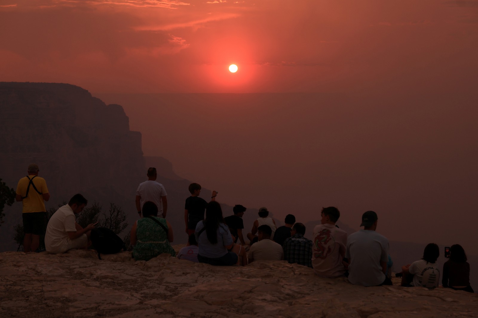 Visitors on the rim of the Grand Canyon watch a deep red sunset through a haze of wildfire smoke.