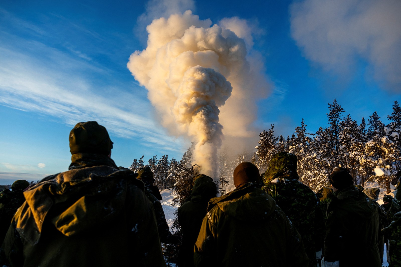 Soldiers watch a signal fire send a plume of smoke up near snow-covered trees.