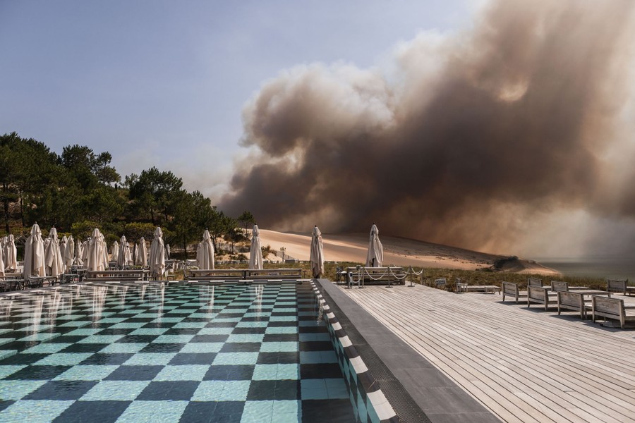 Black smoke rises above a dune, seen from a hotel-swimming-pool deck.