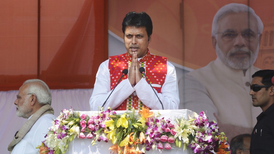 Tripura Chief Minister Biplab Kumar Deb presses his palms together and speaks at a podium adorned with flowers.