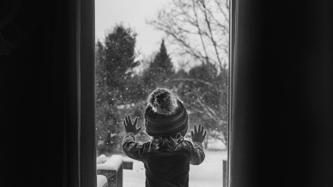 Photo en noir et blanc d'un enfant regardant par la fenêtre alors qu'il neige