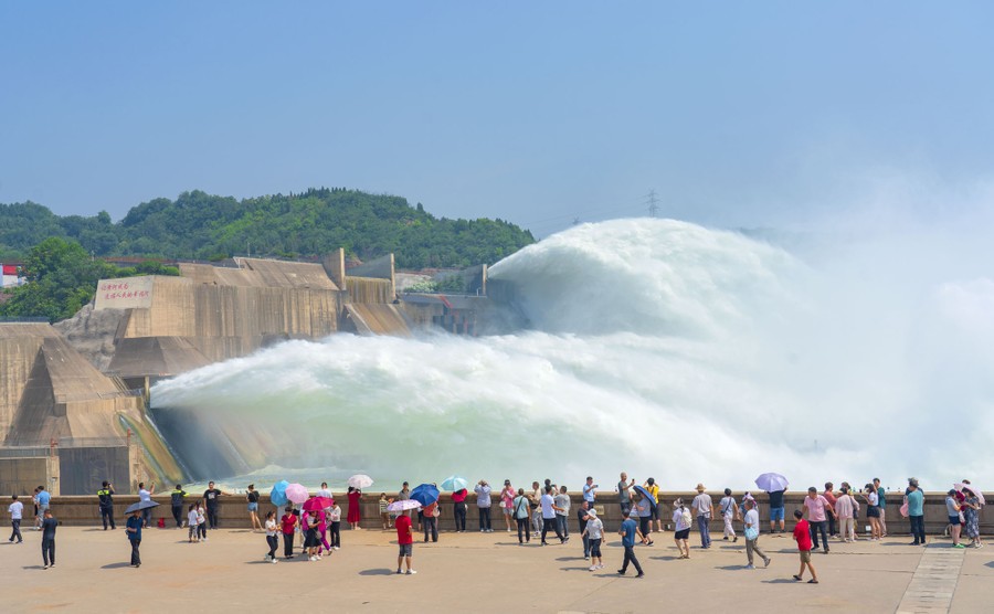 People stand in a viewing area, watching huge plumes of water jetting off the edge of several spillways.
