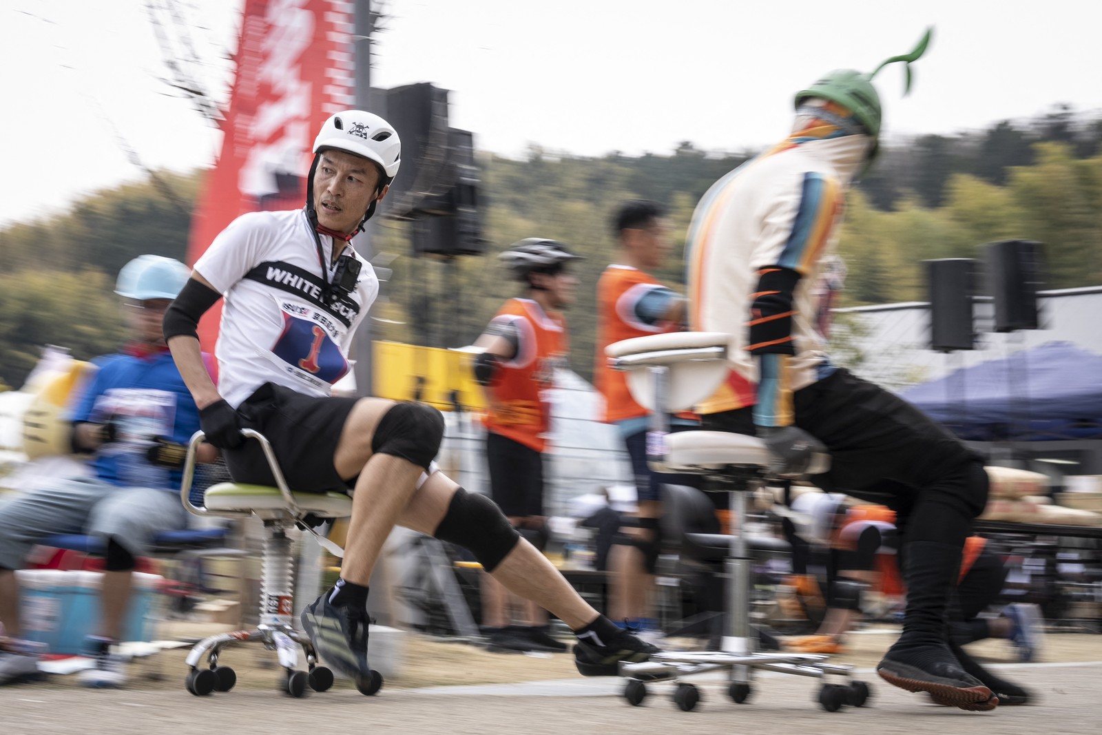 Several people wearing helmets push themselves backwards on wheeled office chairs during an outdoor race.