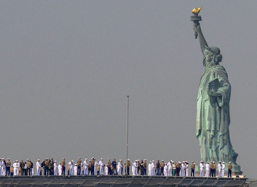 A line of soldiers and sailors stand on a ship's deck as it passes by the Statue of Liberty.