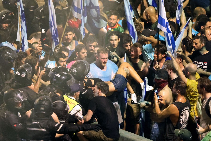A view of a large crowd of protesters facing a group of riot police. A man in the center carries a woman in his arms.