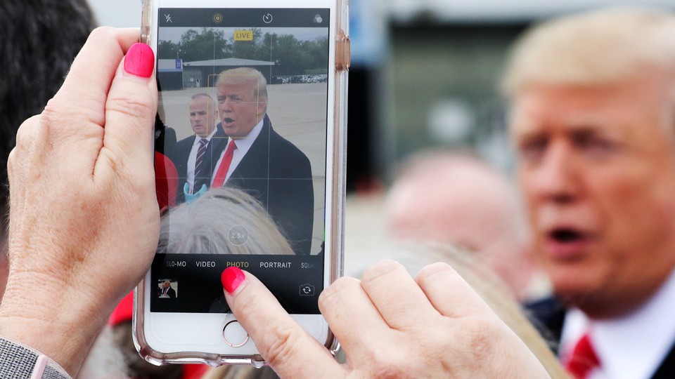 Someone takes a photograph of President Trump on a phone, with Trump visible in the background.