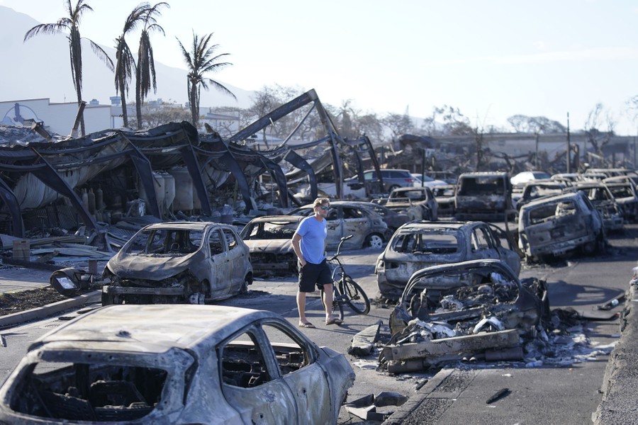 A man stands beside a bicycle among many burned cars and buildings.