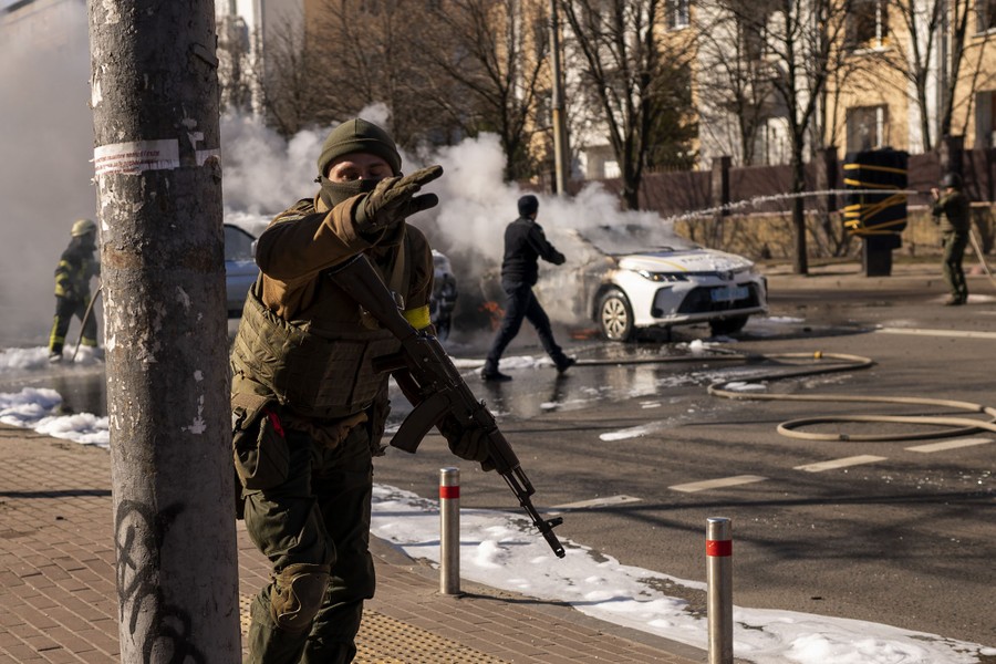 A soldier waves away the photographer as people in the background spray water on several burning cars.