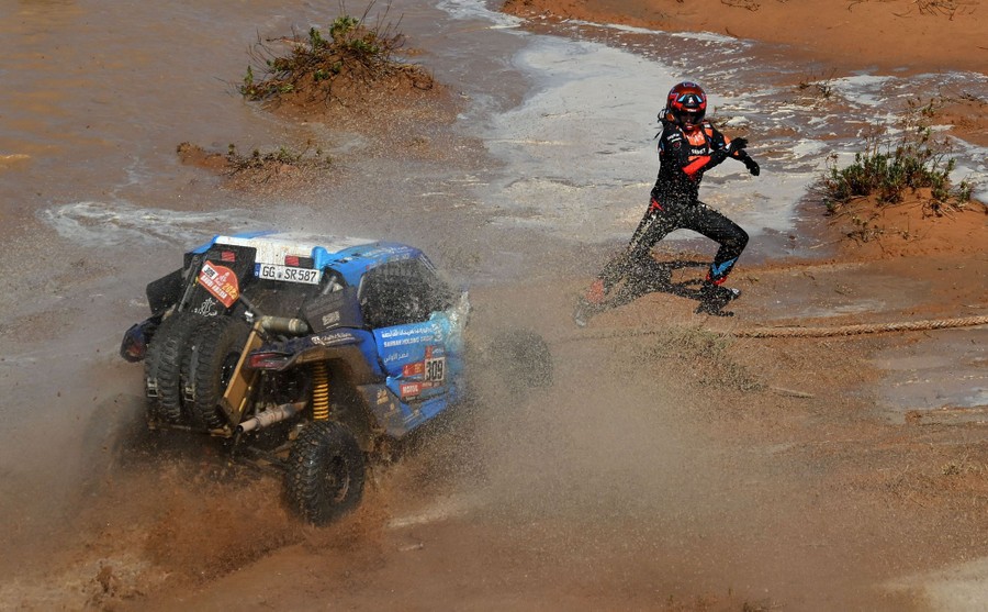A race-car driver (not in a car) steps quickly sideways on a muddy plain, trying to avoid an oncoming race car.