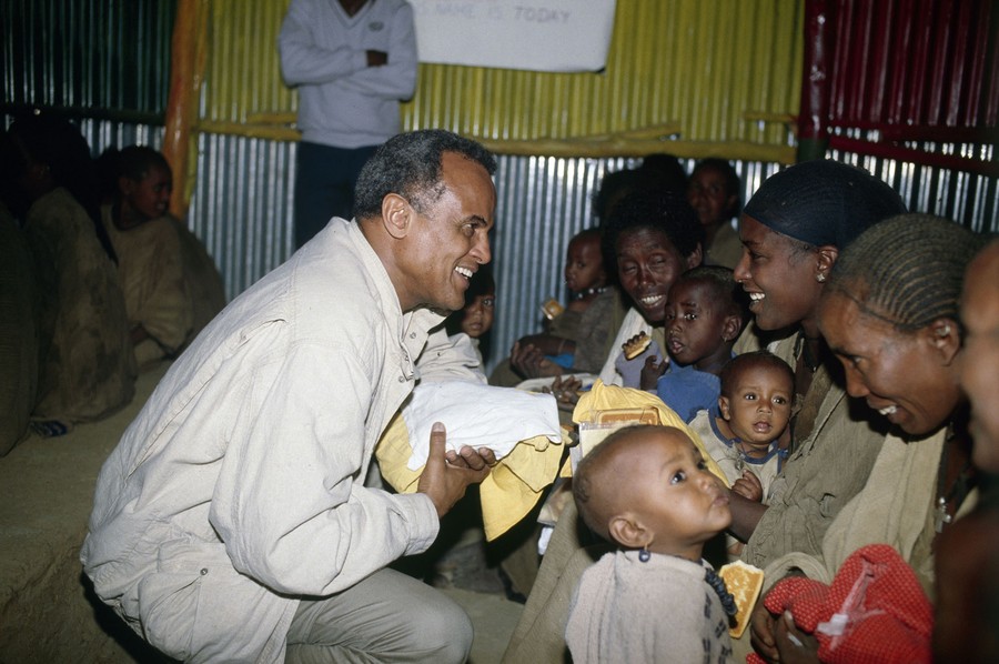 Harry Belafonte kneels, helping distribute food to refugees seated in front of him.