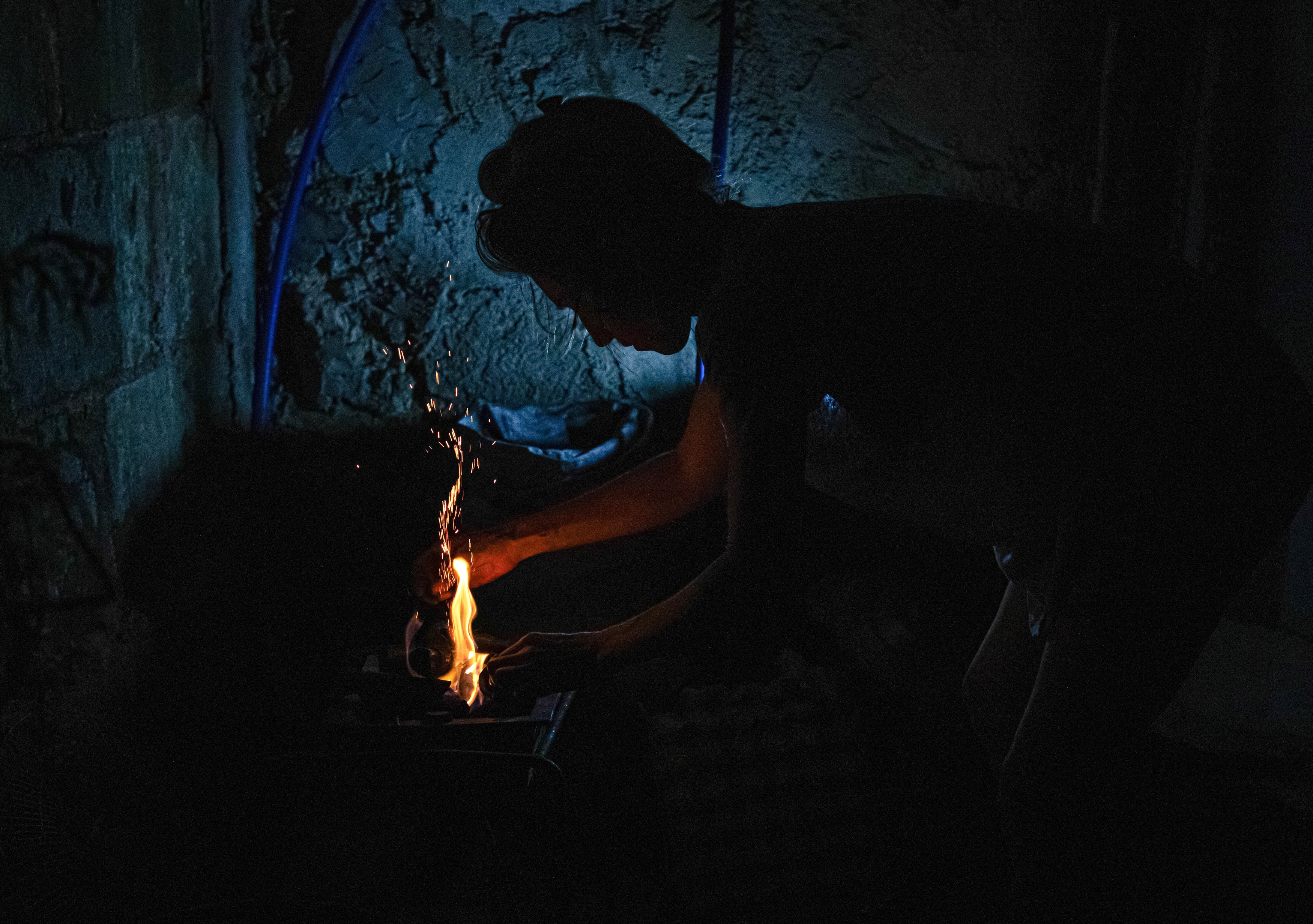 A woman prepares a wood fire to cook food inside a dark room.