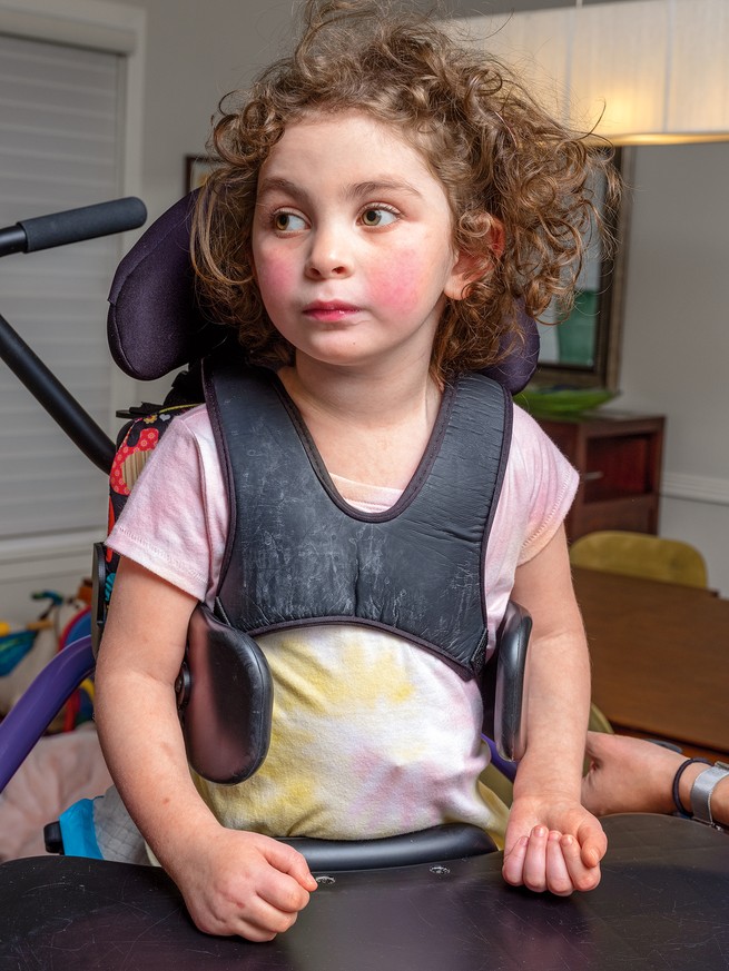 Livie, with curly brown hair and rosy cheeks, looks sideways while standing in her stander, hands resting on a table.
