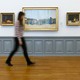 A woman, who is blurry, walks past a series of paintings in a museum.