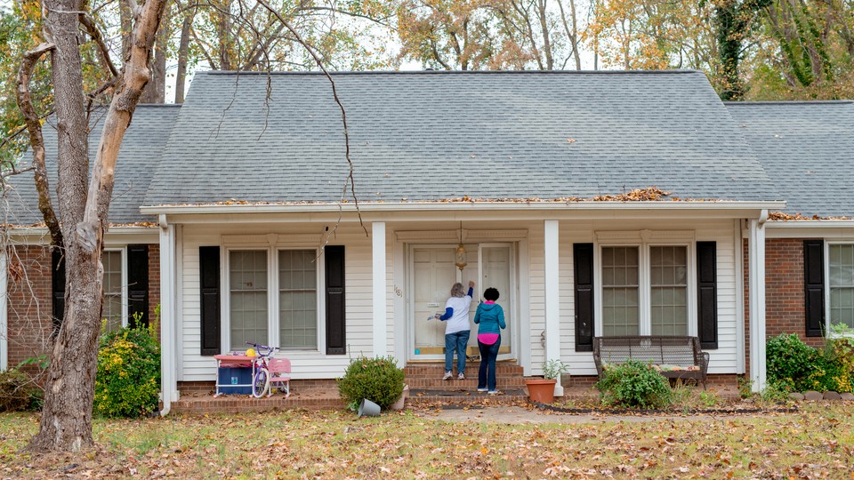 Color photograph of two people knocking on the front door a white and red-brick house with fallen leaves in the yard.