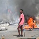 A young man stands in a city street where a fire is burning, emitting a cloud of black smoke.