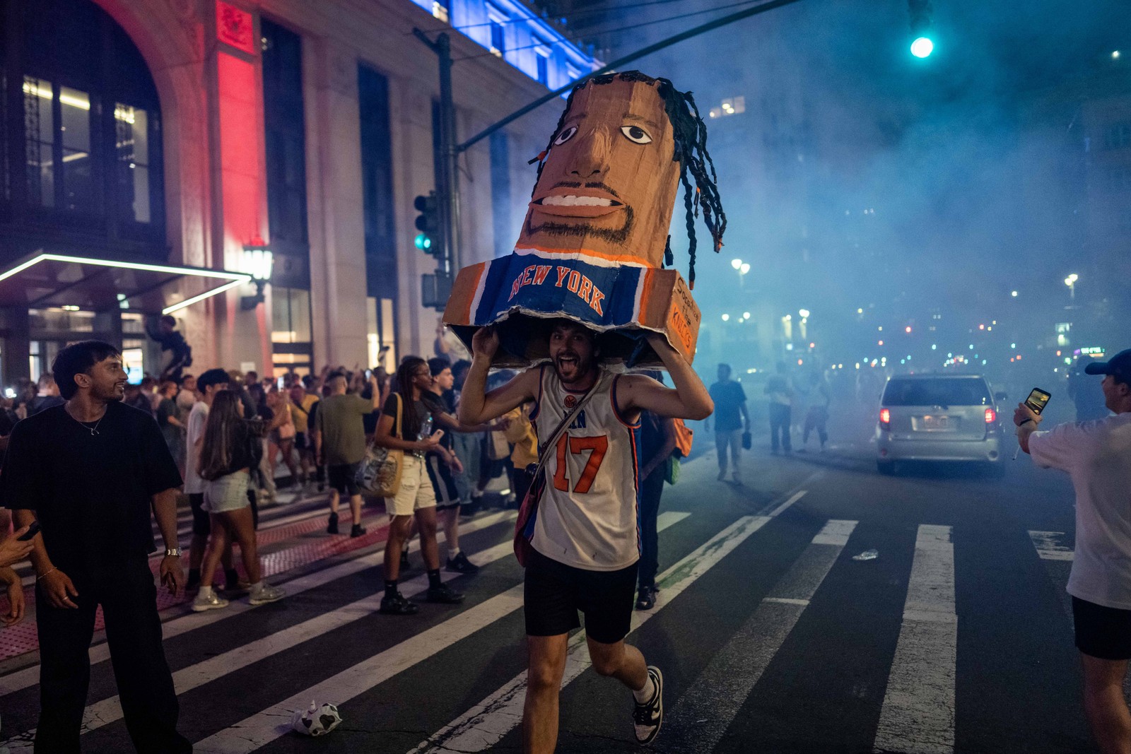 Knicks fans gather in a street, with one carrying a cardboard bust of Jalen Brunson.