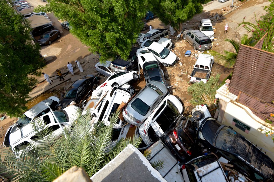 A couple dozen cars sit haphazardly in a pile, amid flood debris, between houses.