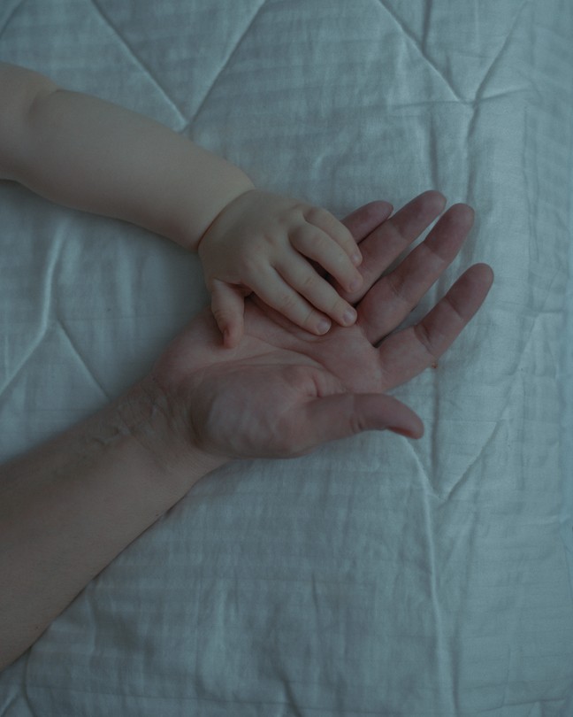 An image of an infant's hand on top of an adult's hand, both resting on a white comforter