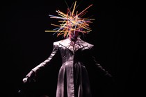 Grace Jones performs onstage wearing a head covering made of many colorful sticks.