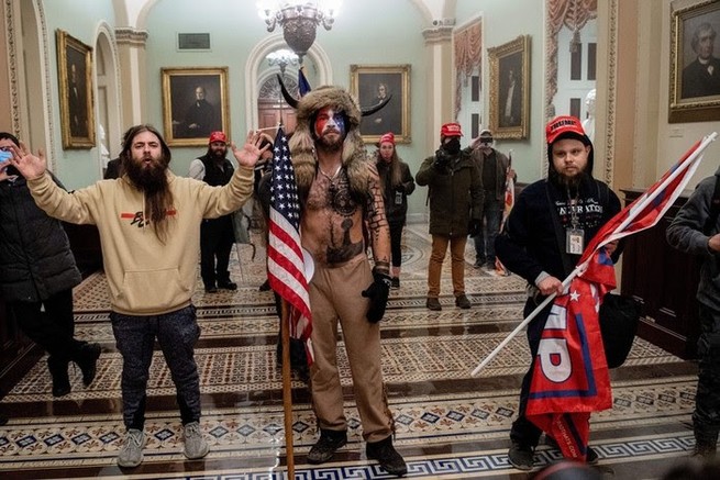 A bare-chested man wearing horns and carrying an American flag stands in the center of this photo taken at the Capitol during the Jan. 6 insurrection. A number of other men, many wearing red baseball caps, are also shown in the photo. 