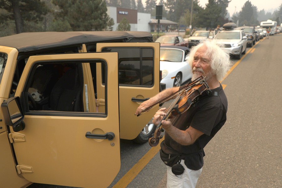 A man stands next to his vehicle playing a violin sitting in a long line of traffic.