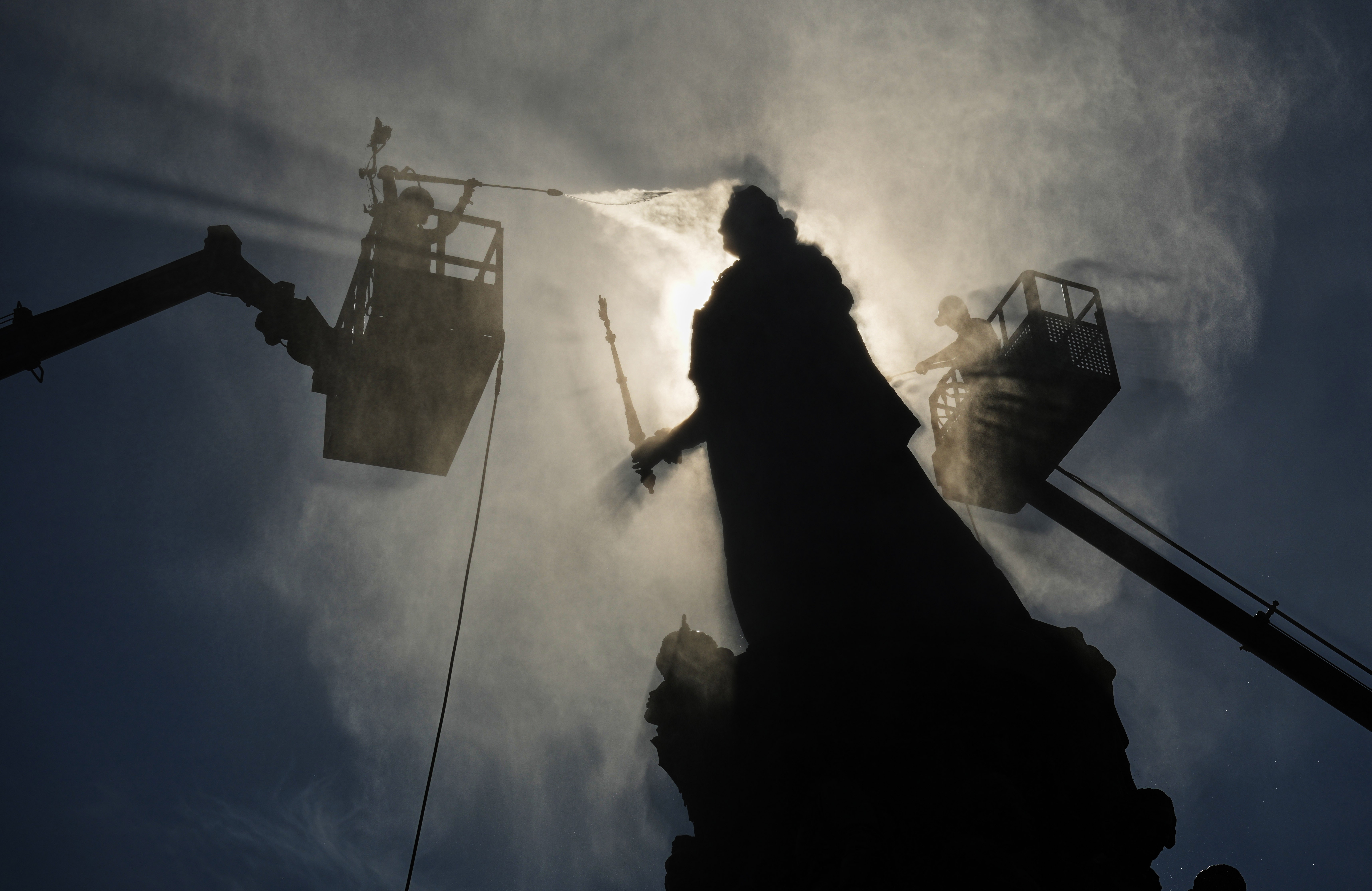 Workers in cherry-pickers spray clean a tall statue.