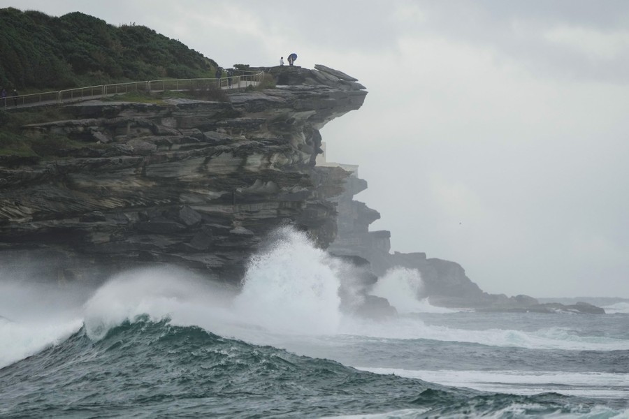 People watch from a cliff top as large waves crash below.