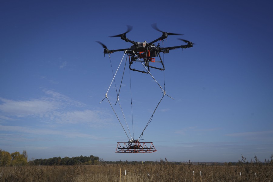 A large drone hovers above a field, carrying equipment to search for mines.