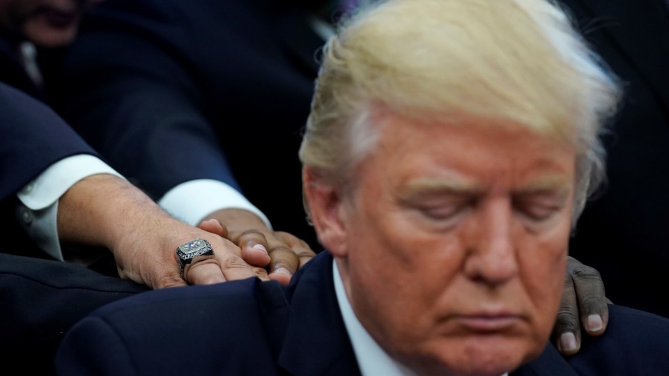 Donald Trump looks down as three men, whose faces are not visible, lay their hands on his back and shoulders.