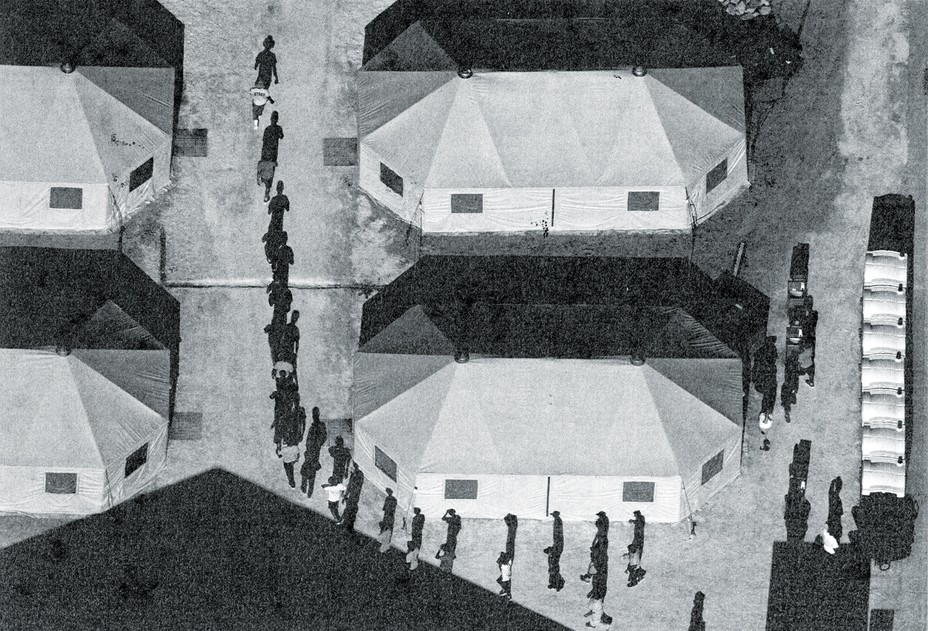 Photo with aerial view of children marching outdoors in the sun casting long shadows in a line around large tents 