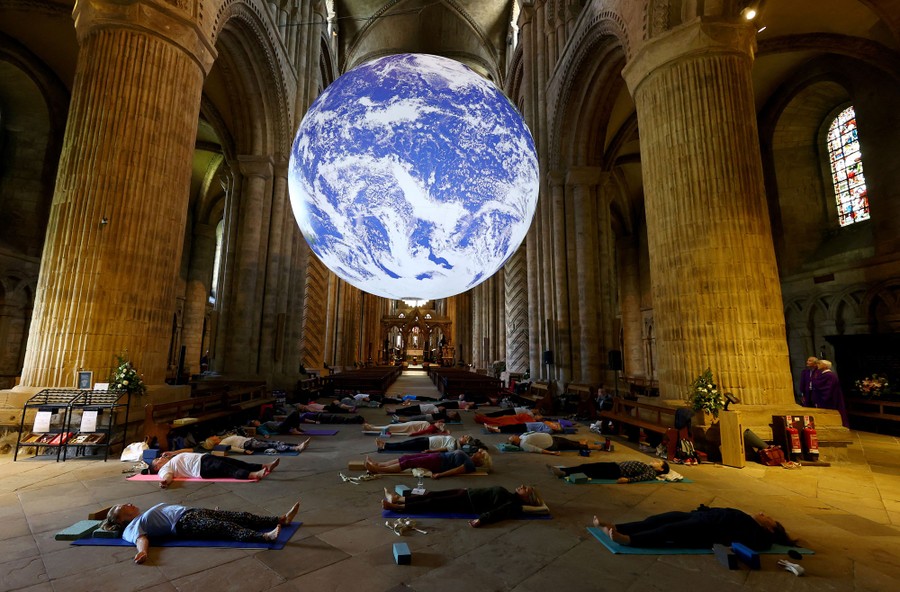 A small group of people attends a yoga class inside a cathedral, directly beneath a large, illuminated sculpture of the Earth.