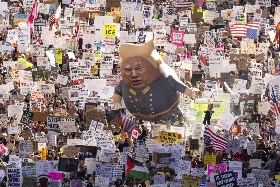 A large crowd of protesters marches down a Los Angeles street carrying many signs and a balloon with an image of President Donald Trump.