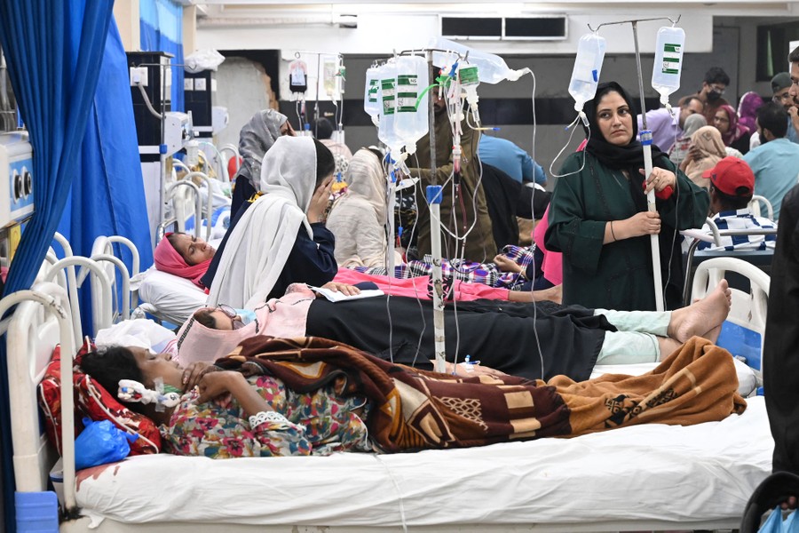 Many people lie on hospital beds and others walk nearby inside a hospital.