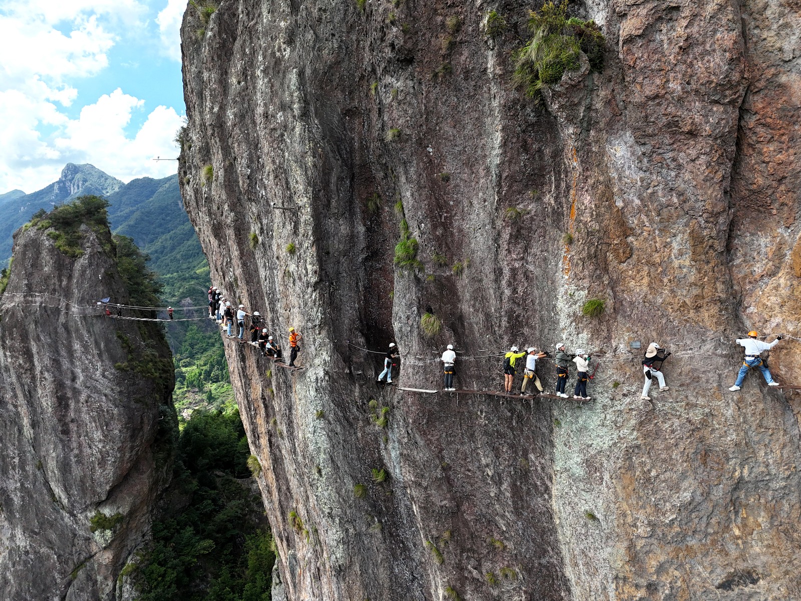Tourists slowly guide themselves along the face of a tall cliff, attached to cables and ropes, walking on narrow planks and footholds.