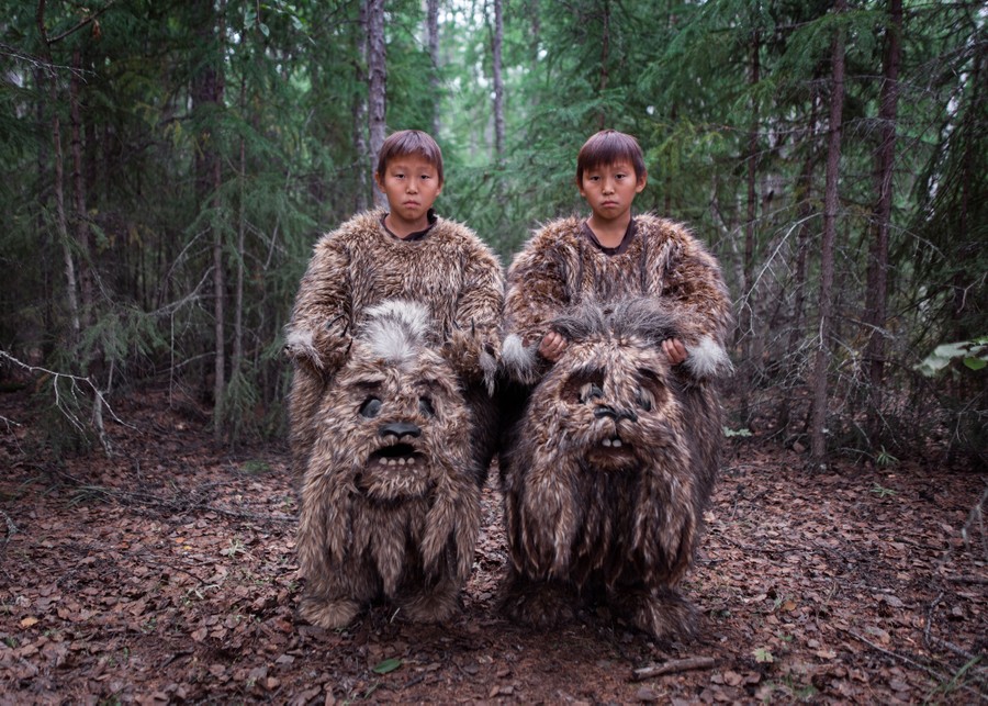 Young twins stand in a forest, wearing shaggy-creature costumes, and holding their costume heads in their hands.
