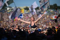 A person sits on top of someone else's shoulders, with their arms held out, in the middle of a large audience at a concert.